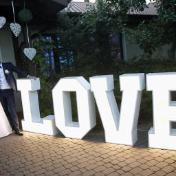 K.M.Usługi Remontowo-Hydrauliczne - Młoda para w strojach ślubnych pozuje obok dużych, podświetlanych liter układających się w słowo LOVE na tle budynku i zieleni.