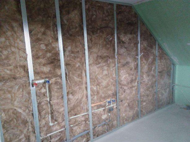 Izolacja ścian wełną mineralną w metalowym stelażu, z widocznymi rurami instalacji wodnej z oznaczeniami ciepłej i zimnej wody, w trakcie prac wykończeniowych poddasza.
