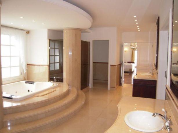 Przestronna łazienka w Warszawie z jacuzzi na podwyższeniu, wykończona beżowym marmurem, widok na umywalkę z chromowaną baterią i lustro.