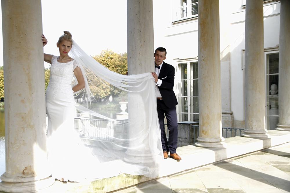 Para młoda w scenerii pałacowej z kolumnami, panna młoda w białej sukni ślubnej i welonie, pan młody w czarnym smokingu, jasne światło słoneczne.