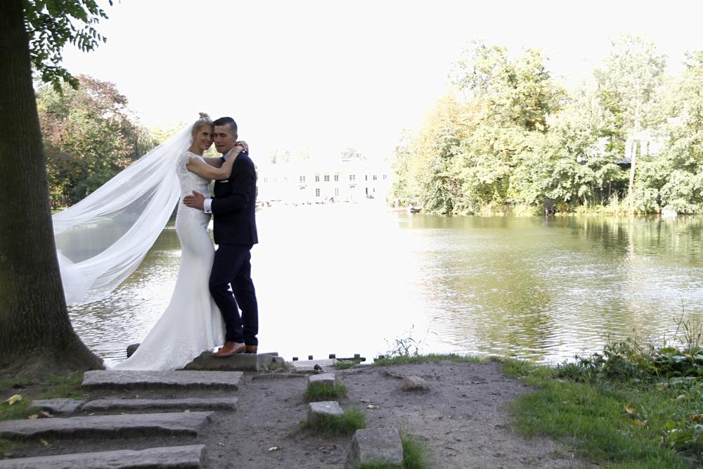 Para młoda w objęciach na tle jeziora i drzew, ona w białej sukni ślubnej z długim welonem, on w granatowym garniturze i brązowych butach, stoją na kamiennych stopniach.
