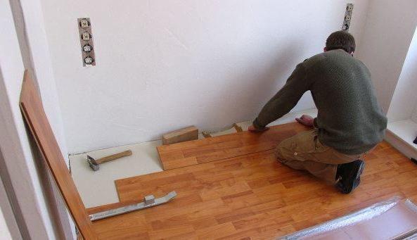 Instalacja paneli podłogowych w rogu pokoju. Widoczne narzędzia, praca w trakcie układania, niezabezpieczone gniazdka elektryczne na ścianie.
