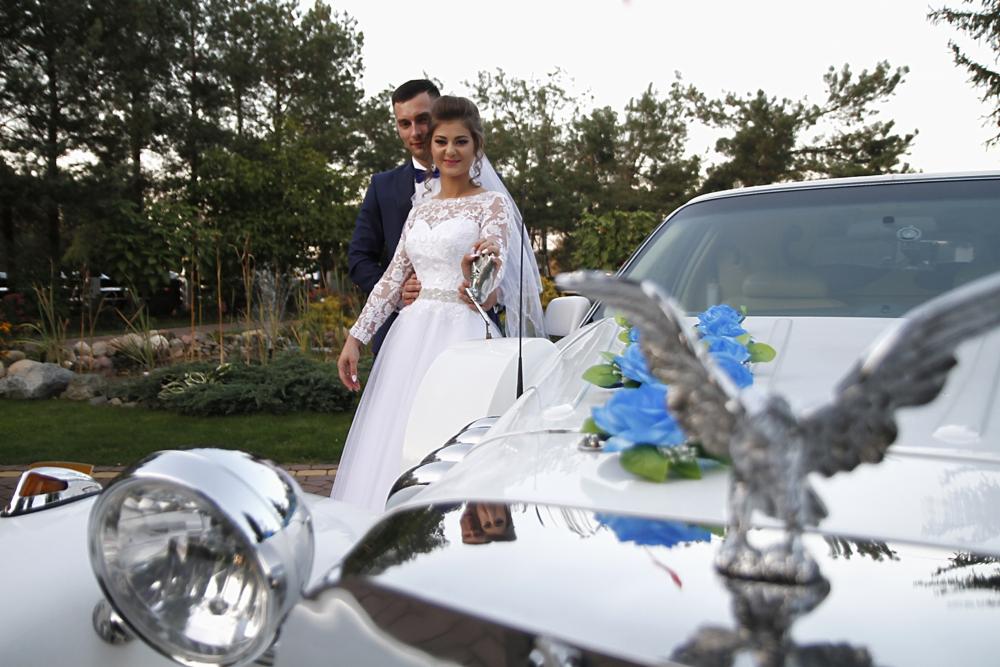 Panna młoda w białej sukni i pan młody w granatowym garniturze pozują obok białej limuzyny z chromowanymi detalami i dekoracją z niebieskich kwiatów oraz figurką orła.