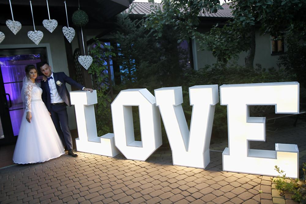 Młoda para w strojach ślubnych pozuje obok dużych, podświetlanych liter układających się w słowo LOVE na tle budynku i zieleni.