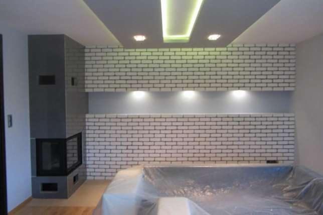 Nowoczesny salon z kominkiem obudowanym szarą konstrukcją i ścianą wyłożoną białą cegłą, oświetloną punktowymi lampami sufitowymi i wnękowymi.
