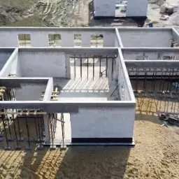 Surowy stan domu z betonu komórkowego w trakcie budowy; widoczne szalunki, podpory i izolacja fundamentów, teren wokół w stanie budowy.