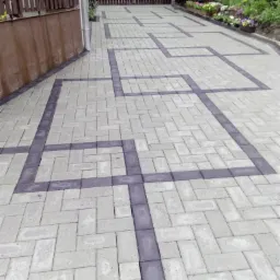 Ułożona kostka brukowa w geometryczny wzór na podwórku, z ciemnoszarymi obramowaniami tworzącymi kwadraty połączone liniami, widok na drewniany płot i rabatę kwiatową w tle.