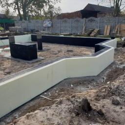 Usługi Budowlane Electro-Bud - Ocieplone fundamenty domu jednorodzinnego w trakcie budowy, widoczne czarne hydroizolacje i biały styropian, pracownik docinający izolację. W tle ogrodzenie z prefabrykatów betonowych i zabudowa...