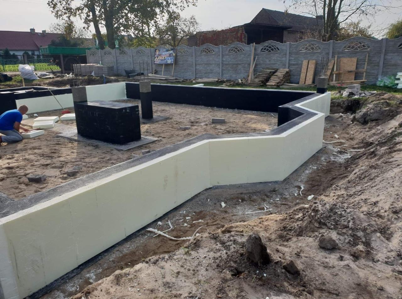 Ocieplone fundamenty domu jednorodzinnego w trakcie budowy, widoczne czarne hydroizolacje i biały styropian, pracownik docinający izolację. W tle ogrodzenie z prefabrykatów betonowych i zabudowa...