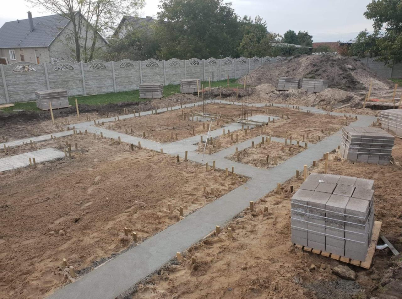 Wylane betonowe ławy fundamentowe z drewnianymi kołkami, zbrojenie wystające w górę, ułożone palety z kostką brukową w tle, betonowy płot panelowy.