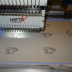 Automatyczna hafciarka nanosi wzór filiżanek na jasny materiał, widoczne kontury kolejnych wzorów.