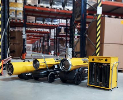 Trzy żółte nagrzewnice MASTER ustawione w magazynie z wysokimi regałami wypełnionymi kartonami, widok z niskiej perspektywy.