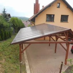 Zadaszenie drewniane z brązowym, lekko przezroczystym dachem przylegające do domu w kolorze piaskowym, z widocznymi samochodami w tle.