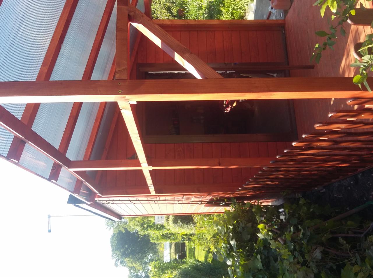 Konstrukcja z drewna i przezroczystego poliwęglanu, prawdopodobnie zadaszenie, z widocznymi elementami mocującymi, pomalowana na czerwono-brązowy kolor, z fragmentem ażurowego ogrodzenia i zieleni...