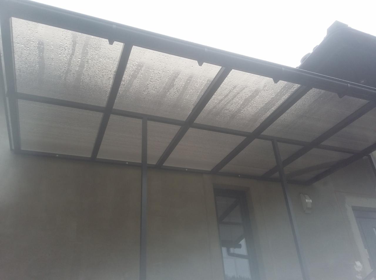 Zadaszenie tarasu z poliwęglanu na metalowej konstrukcji, widoczne krople deszczu na powierzchni, betonowa ściana budynku w tle.