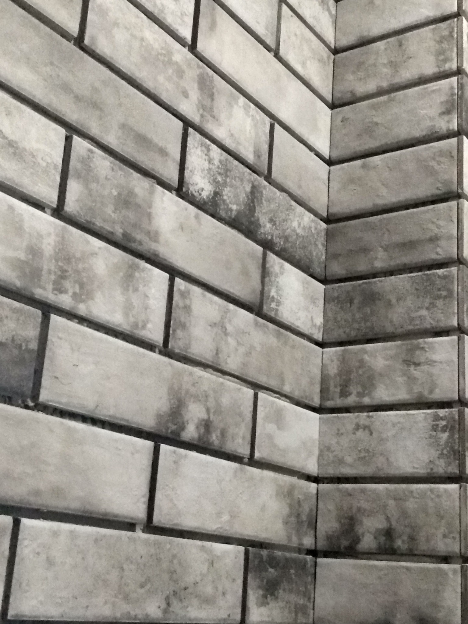 Szara, matowa okładzina ścienna imitująca cegłę, ułożona pod kątem w rogu pomieszczenia z widocznymi ciemniejszymi przebarwieniami.