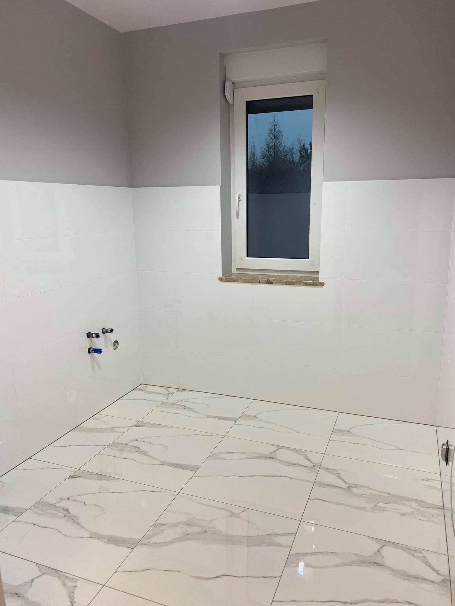 Wykończona łazienka z białymi płytkami imitującymi marmur na podłodze i ścianach do połowy wysokości, widoczne przyłącza hydrauliczne i okno z widokiem na drzewa.