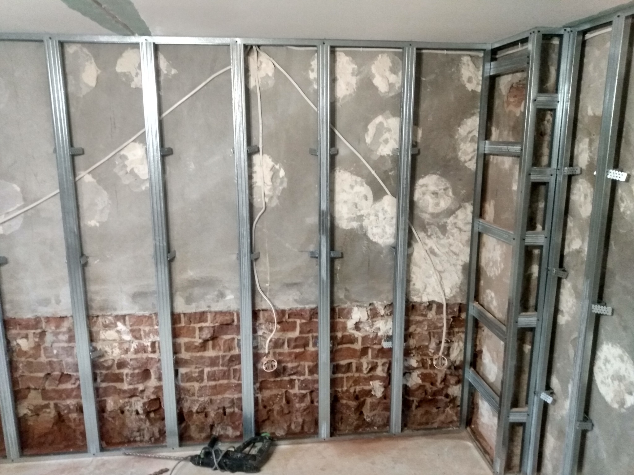 Metalowy szkielet konstrukcji ścianki działowej z płyt gipsowo-kartonowych, widoczne przewody elektryczne i fragment ściany z cegły, w tle ściana z plamami po szpachlowaniu.