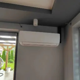 Biały klimatyzator ścienny zamontowany w szarym pomieszczeniu z widokiem na ogród przez okno.