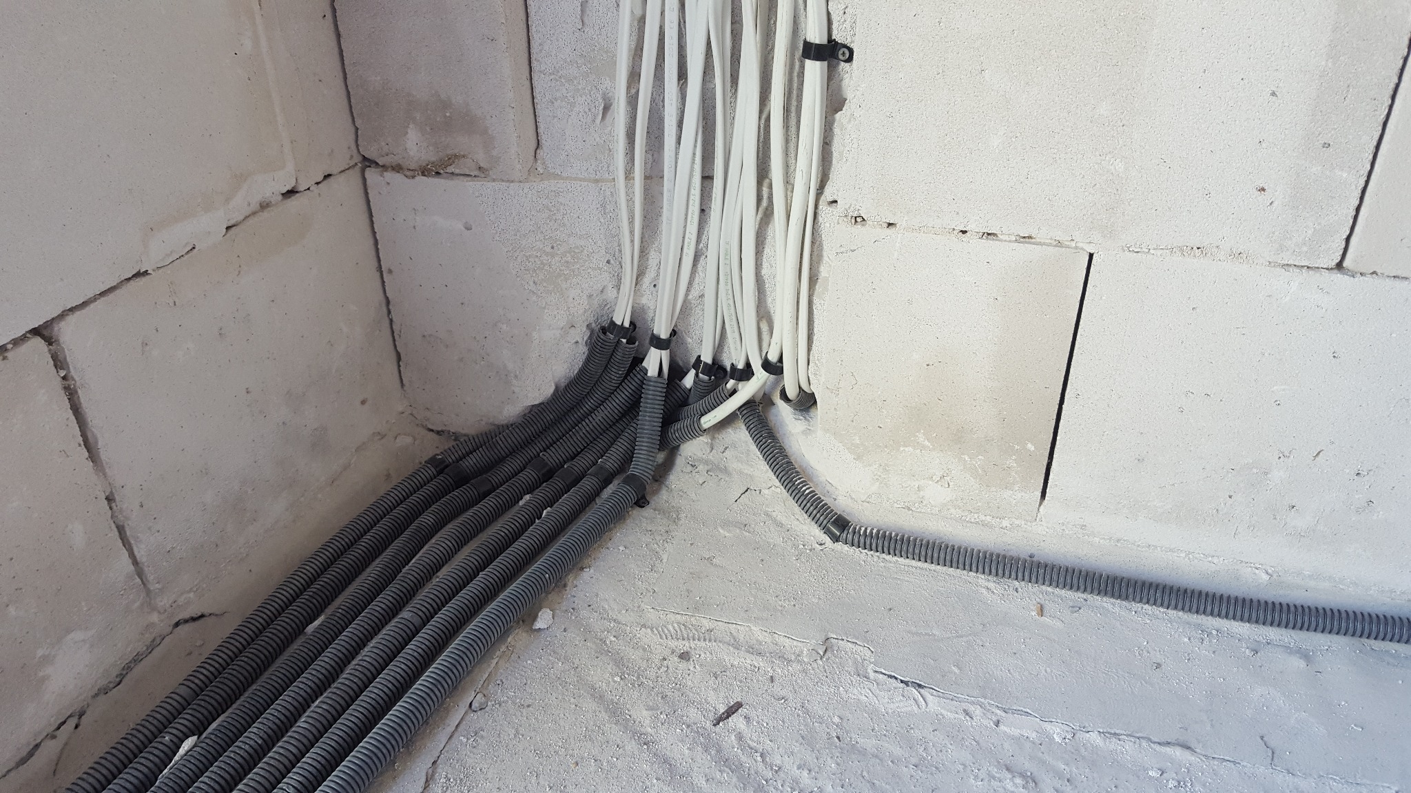 Wiązka białych przewodów elektrycznych wychodzących ze ściany z szarego betonu, zabezpieczonych czarnymi opaskami zaciskowymi, przechodzących w szare, karbowane rury ochronne ułożone na podłodze...