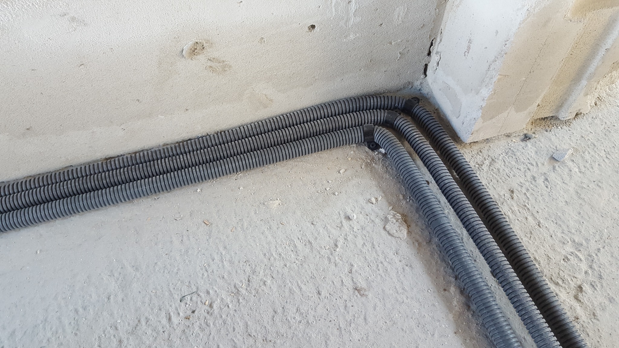 Trzy ciemnoszare karbowane rury prowadzone wzdłuż szarej betonowej ściany i podłogi w surowym stanie.