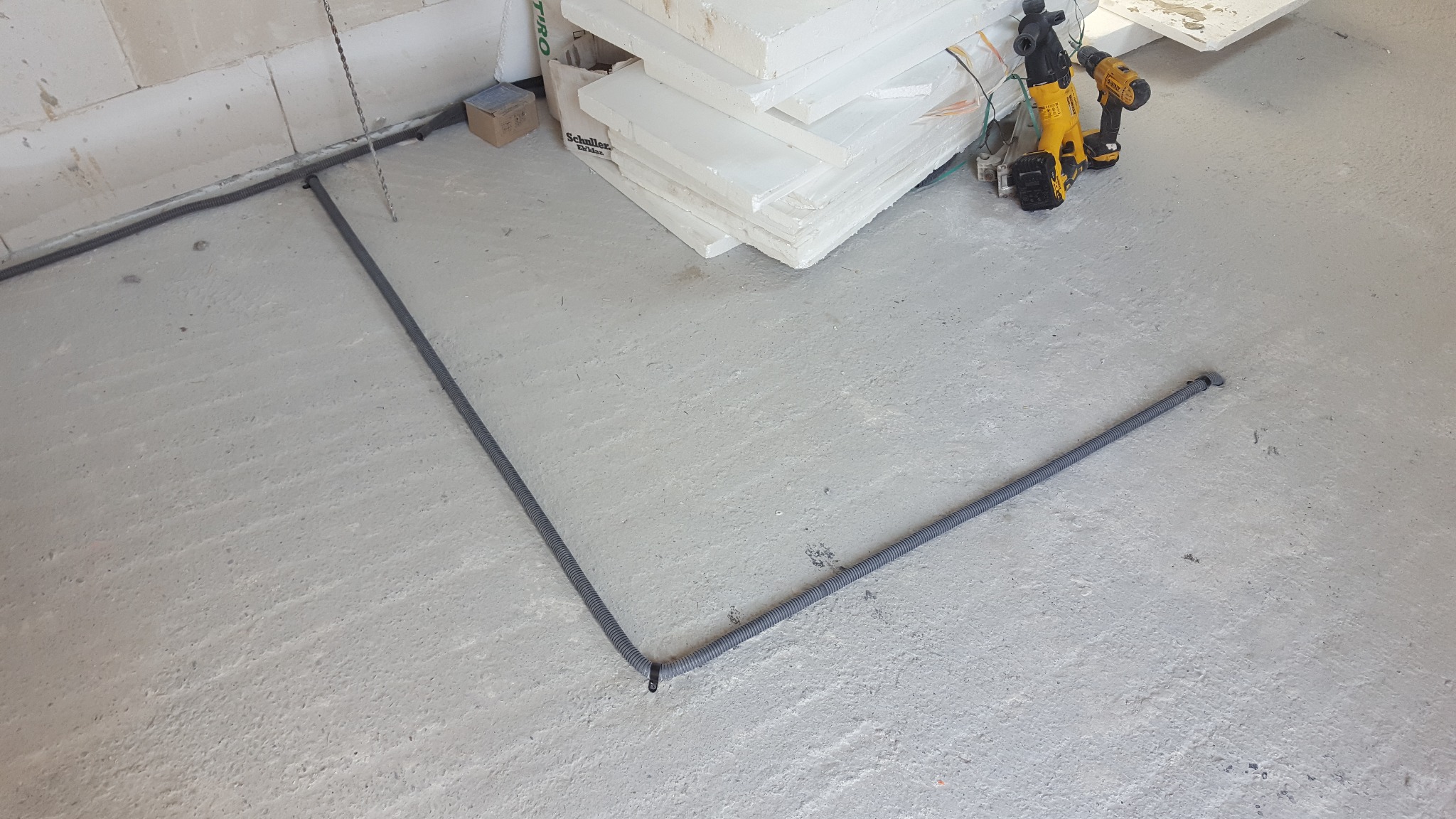 Szara, karbowana rura ochronna ułożona pod kątem prostym na surowym betonowym podłożu wewnątrz budynku, w tle narzędzia DeWalt i styropian.
