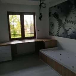 Pokój młodzieżowy z biurkiem narożnym zintegrowanym z parapetem, szafką i łóżkiem, na ścianie fototapeta z motywem zębatek.