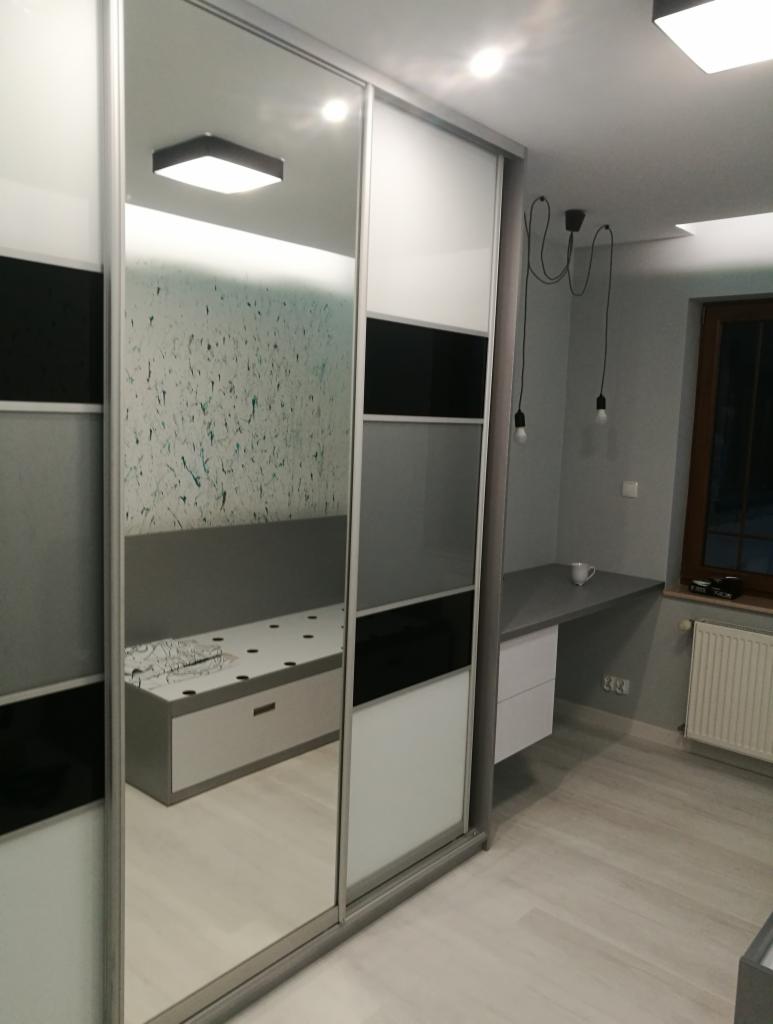 Nowoczesna szafa przesuwna z lustrem, z panelami w kolorze białym, szarym i czarnym, z widocznym biurkiem i lampą wiszącą w tle.