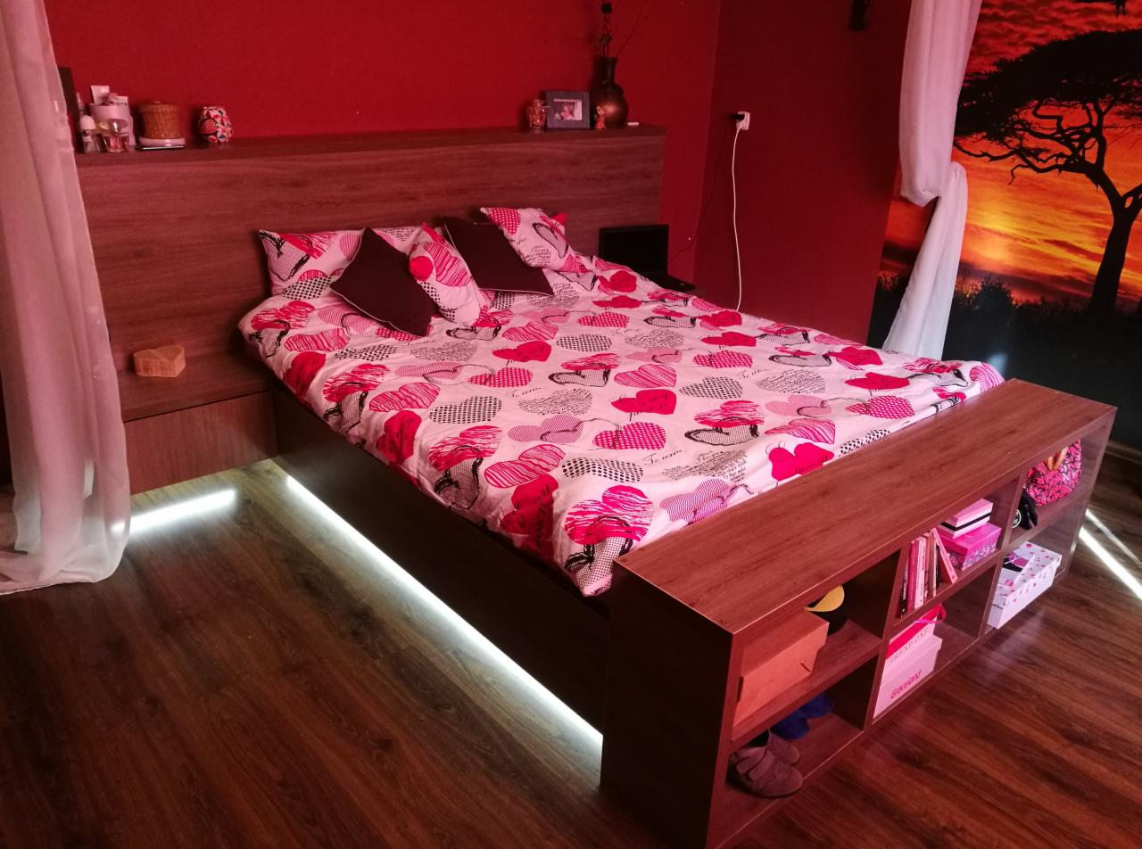 Drewniane łóżko z podświetleniem LED, zabudowane wezgłowie z półkami i szafkami nocnymi, pościel w serca, czerwone ściany i dekoracja afrykańska.