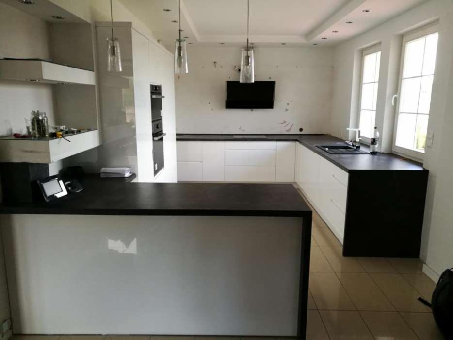 Wnętrze kuchni w trakcie montażu: białe szafki z czarnym blatem, wyspa kuchenna, wiszące lampy, ślady po pracach na ścianie.