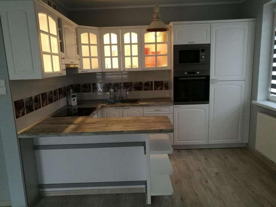 Biała kuchnia z drewnianym blatem i wyspą, górne szafki z przeszklonymi frontami, wbudowany piekarnik i mikrofalówka, dekoracyjne płytki nad blatem, podłoga imitująca drewno.