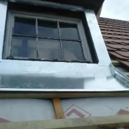 Obróbka blacharska okna dachowego z widocznymi nitami mocującymi, na tle częściowo pokrytego dachówką dachu.