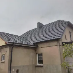 Dom jednorodzinny z nowym, grafitowym dachem z blachodachówki i ciemnymi rynnami, widok z boku na tle pochmurnego nieba.