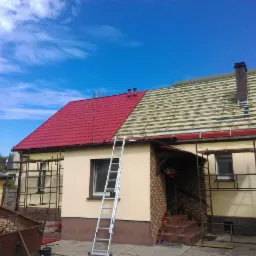 Dom z częściowo wymienionym, czerwonym dachem z blachy trapezowej, widoczne rusztowania i drabina oparta o ścianę, przygotowane łatowanie pod resztę pokrycia.