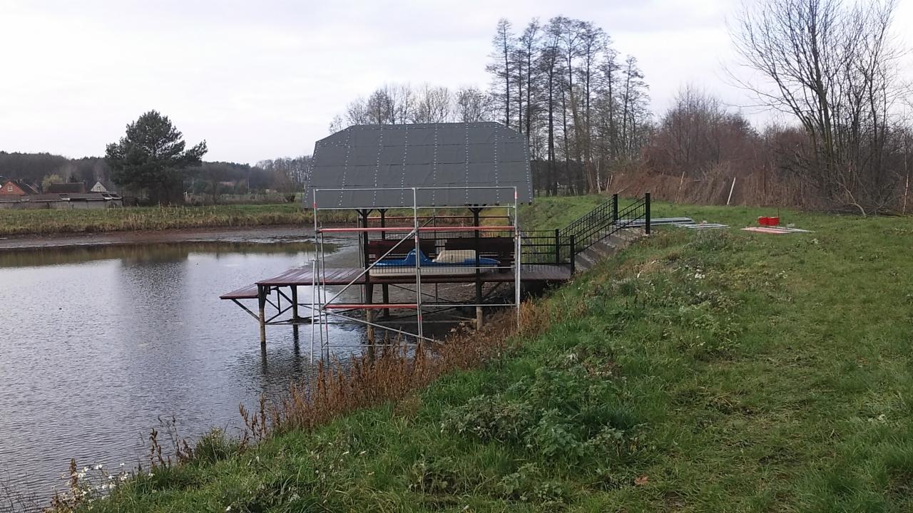 Pomost nad wodą z zadaszeniem w trakcie budowy, widoczne rusztowanie i dach z blachy trapezowej na tle drzew i pochmurnego nieba.