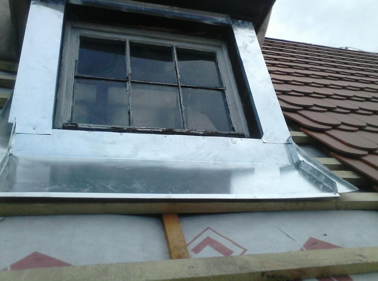 Obróbka blacharska okna dachowego z widocznymi nitami mocującymi, na tle częściowo pokrytego dachówką dachu.