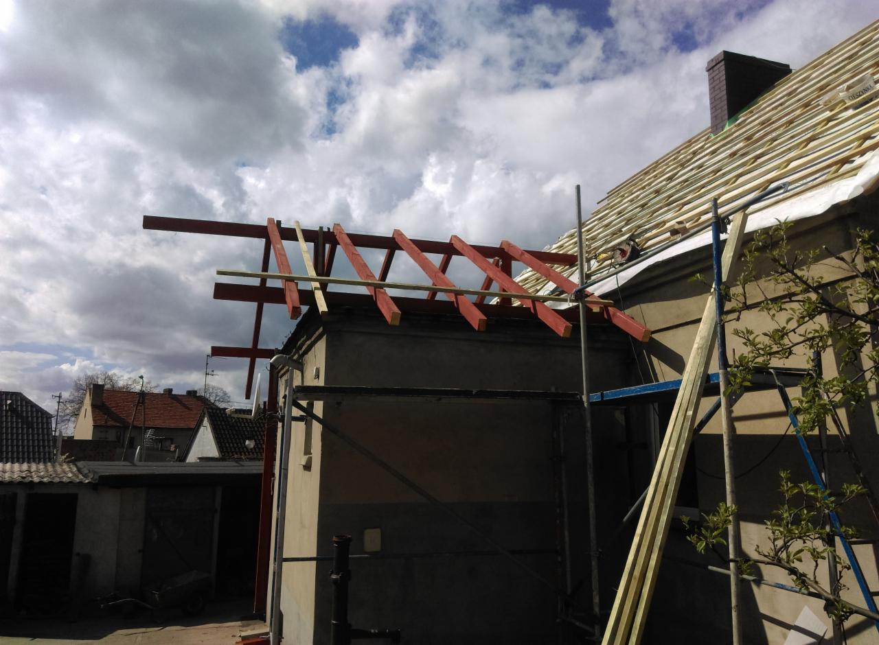 Konstrukcja więźby dachowej z czerwonymi elementami, widok z dołu na budynek z częściowo ułożonymi łatami, w tle pochmurne niebo i sąsiednie domy.