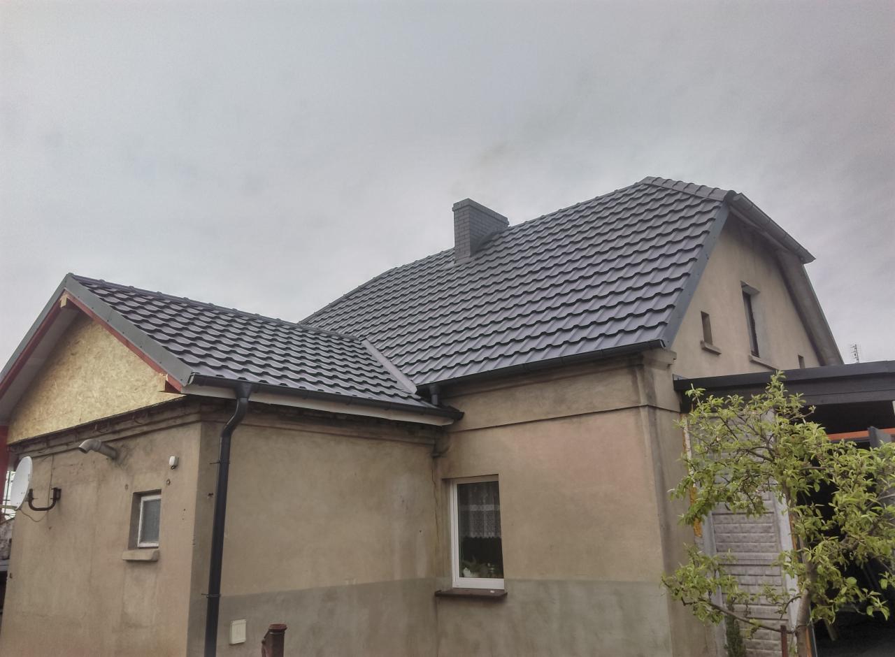 Dom jednorodzinny z nowym, grafitowym dachem z blachodachówki i ciemnymi rynnami, widok z boku na tle pochmurnego nieba.