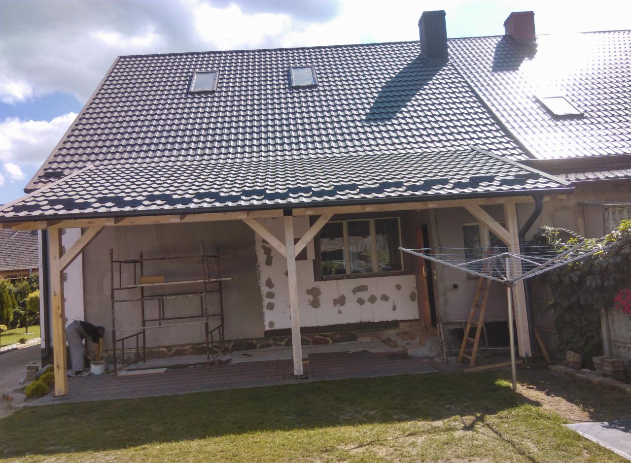 Dom z grafitowym dachem z dachówki, widoczne okna dachowe i komin, dobudowany drewniany ganek w trakcie wykończenia, rusztowanie i pracownik przy pracy.