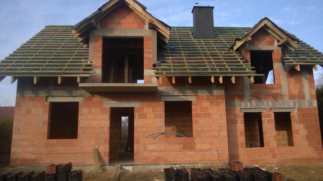 Surowy stan domu jednorodzinnego w trakcie budowy, widoczna konstrukcja więźby dachowej z łatami, komin oraz stosy dachówek na ziemi.