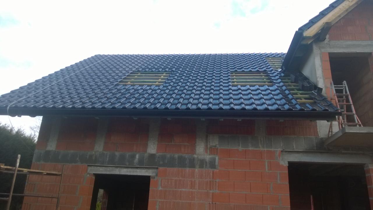 Nowy dach z ciemnej dachówki ceramicznej na surowym budynku z czerwonej cegły, widoczne okna dachowe w trakcie montażu i rusztowanie.