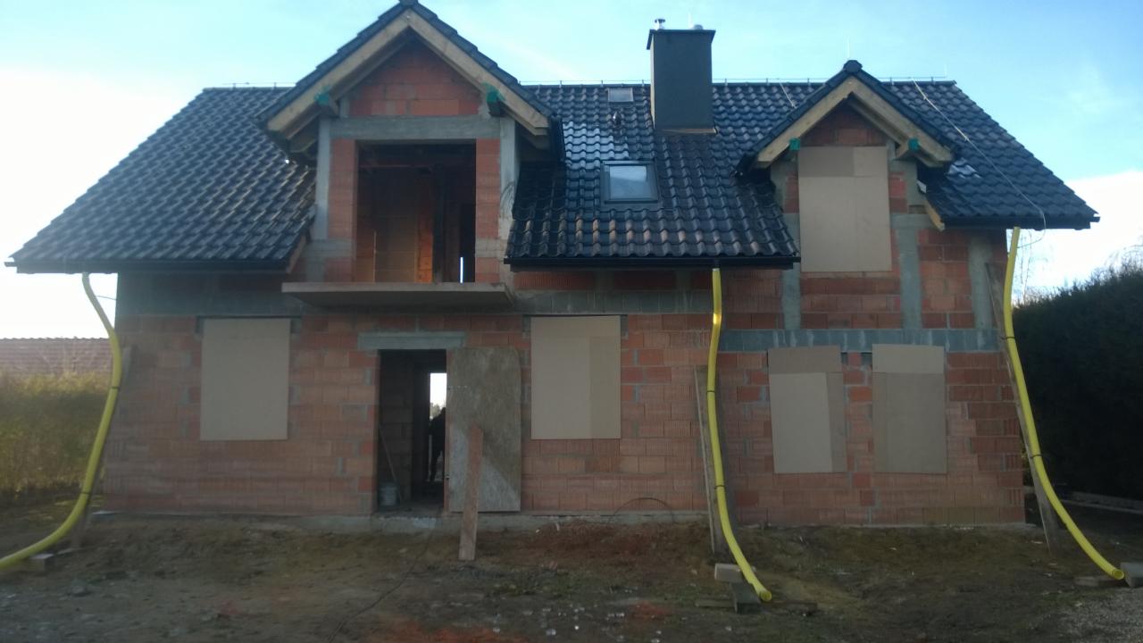 Niedokończony dom z cegły z czarną dachówką, widoczne rynny i okna zabite płytami, żółte rury na elewacji.