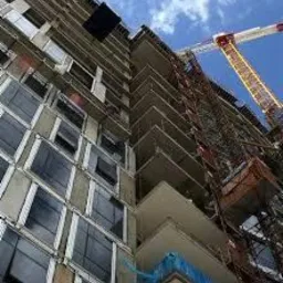 Wysoki budynek w trakcie budowy, widok z dołu, z widocznym żurawiem budowlanym na tle błękitnego nieba z chmurami. Konstrukcja w stanie surowym, z licznymi oknami i balkonami.