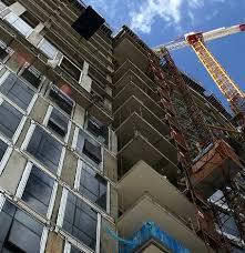 Wysoki budynek w trakcie budowy, widok z dołu, z widocznym żurawiem budowlanym na tle błękitnego nieba z chmurami. Konstrukcja w stanie surowym, z licznymi oknami i balkonami.