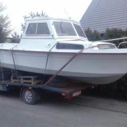 Waldemaro - Biała łódź motorowa zabezpieczona pasami na platformie białego busa, widoczne drewniane palety pod łodzią, tło stanowią dom i krzewy.