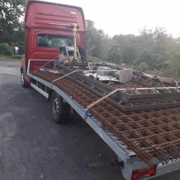 Waldemaro - Czerwony bus z platformą transportową przewozi siatkę zbrojeniową i ubijarkę wibracyjną na tle zieleni.