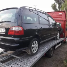 Czarny samochód Ford Galaxy kombi z bagażnikiem dachowym, z widoczną tablicą rejestracyjną 96YR, znajduje się na metalowej platformie lawety koloru srebrnego, która jest przymocowana do czerwonego...
