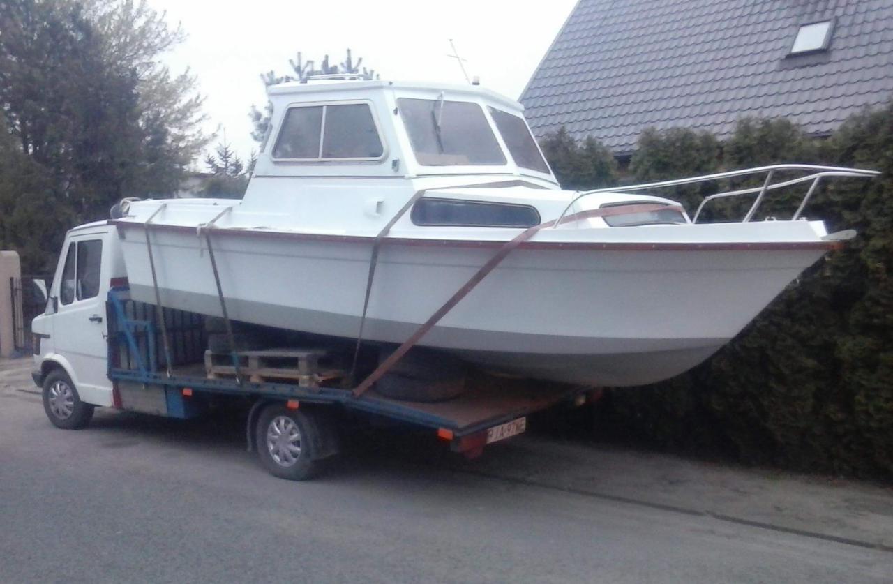 Biała łódź motorowa zabezpieczona pasami na platformie białego busa, widoczne drewniane palety pod łodzią, tło stanowią dom i krzewy.