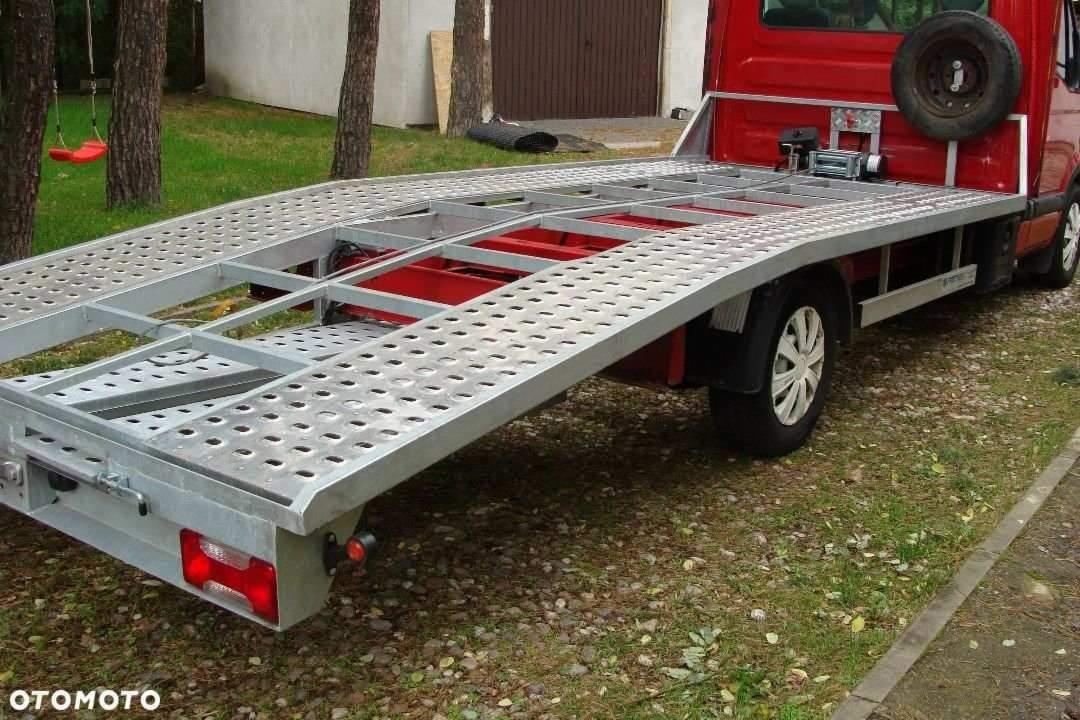 Czerwony samochód z platformą lawety o perforowanej powierzchni, widoczna wyciągarka i zapasowe koło z tyłu kabiny, na trawiastym podłożu.