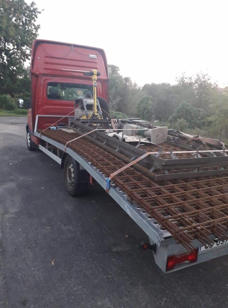 Czerwony bus z platformą transportową przewozi siatkę zbrojeniową i ubijarkę wibracyjną na tle zieleni.